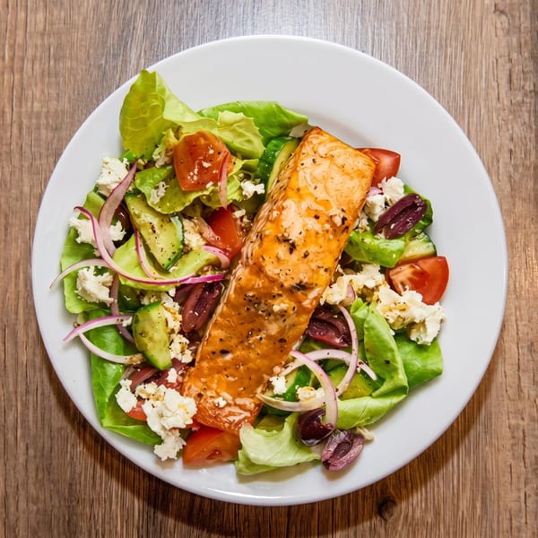 A grilled salmon fillet on a fresh green salad with feta, olives, tomatoes, and red onion.