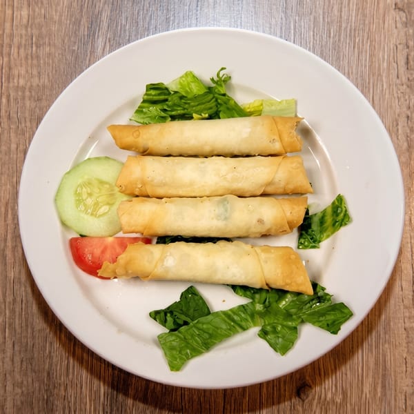 Four golden-brown spring rolls with lettuce, cucumber, and tomato on a white plate.