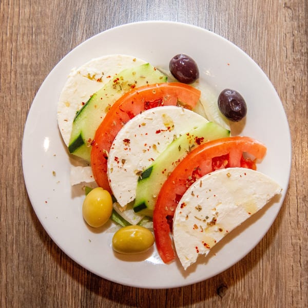 A white plate with sliced feta cheese, tomatoes, cucumbers, and olives on a wooden table.