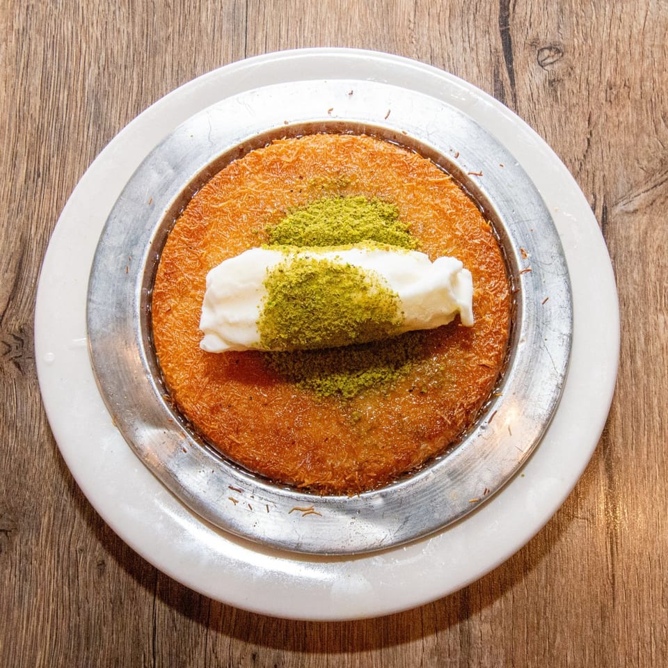 A golden kunefe dessert, topped with clotted cream and green pistachio powder, on a white plate.