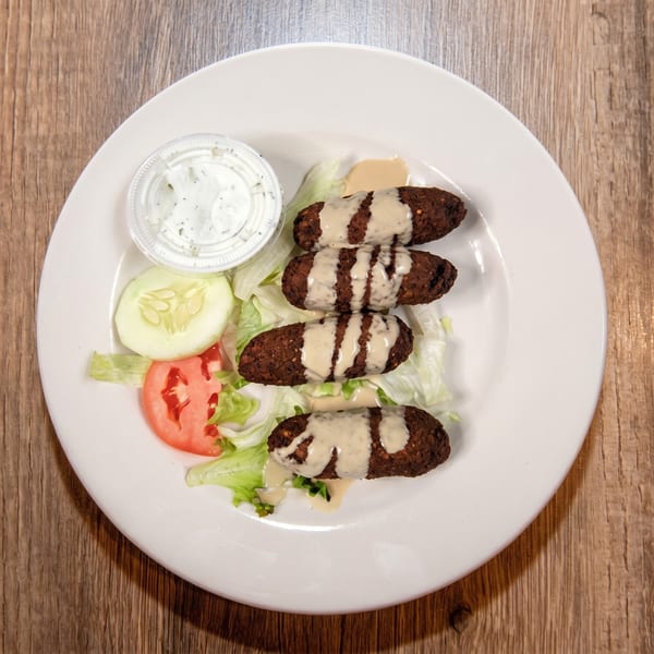 Four falafel pieces drizzled with sauce, served on lettuce with cucumber, tomato, and a side of dip.