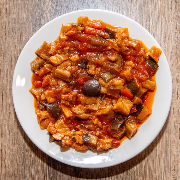 Eggplant caponata with a single black olive on a white plate, on a wooden table.