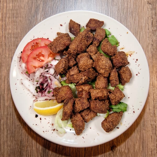 A white plate of crispy fried liver pieces with sliced tomatoes, onions, and a lemon wedge.