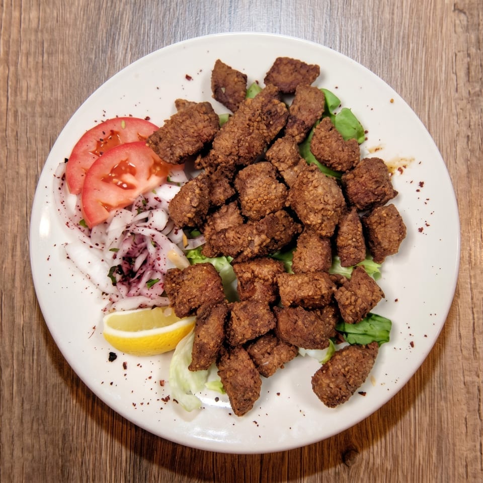 A white plate of crispy fried liver pieces with sliced tomatoes, onions, and a lemon wedge.