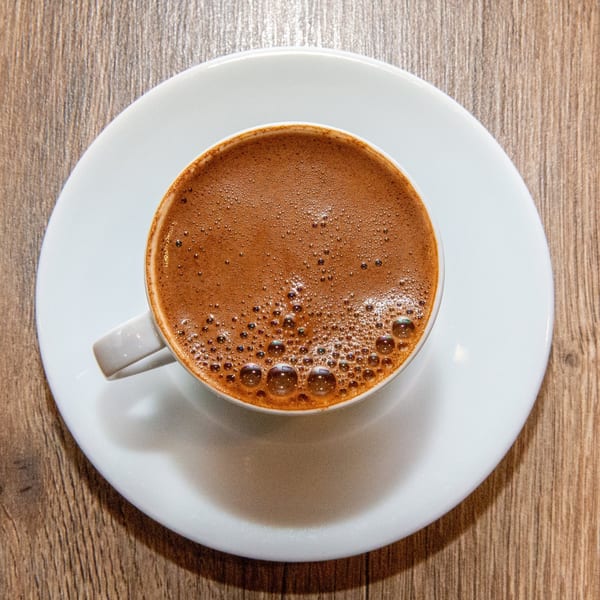Overhead view of a white cup of frothy Turkish coffee on a matching saucer on a wooden table.