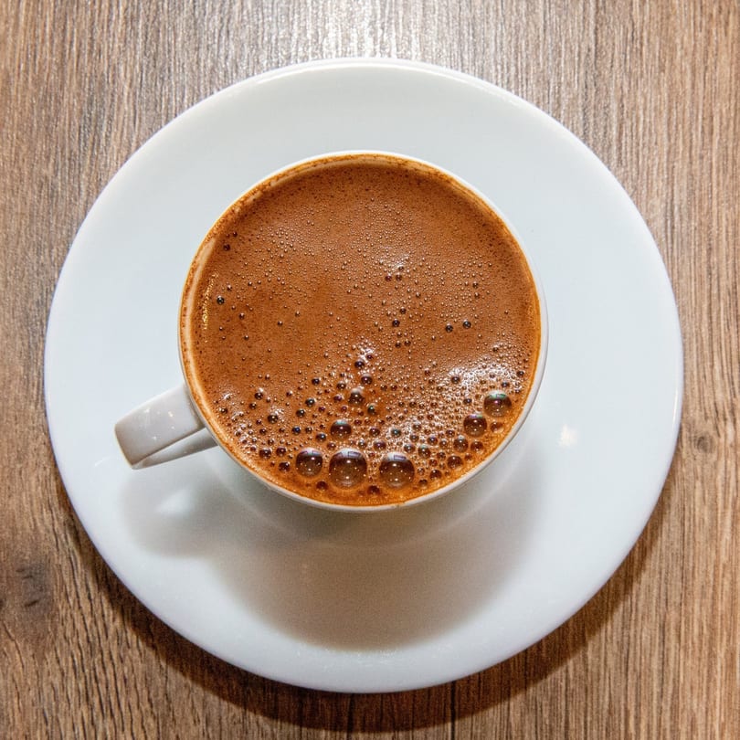 Overhead view of a white cup of frothy Turkish coffee on a matching saucer on a wooden table.